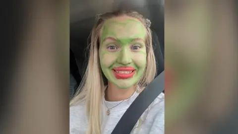 A woman with long blonde hair has painted her face bright green and has bright red lipstick on. She is smiling at the camera. 