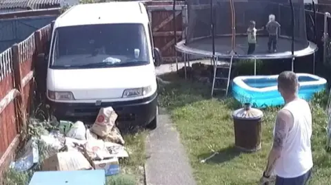 North Wales Police/PA Media A garden with a white van parked on one side and piles of rubbish in front of it. On the other side is a patch of grass with a trampoline and a paddling pool and a black rubbish bin. On the trampoline are two young children. The face of one of the children has been blurred. Stood at the front of the shot, with his back to the camera, is a man with brown hair wearing a white vest and dark trousers.
