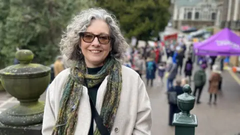 Georgia Roberts/BBC Jane Gill outside the council HQ. She has curly, white hair and glasses. She is wearing a green and yellow scarf and a cream coat. 