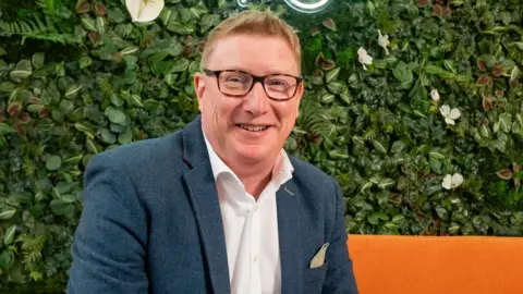 Matt Travis A man with short auburn hair is wearing glasses, a grey suit jacket and a white open neck shirt. He is sitting on a bright orange sofa in front of a green bush