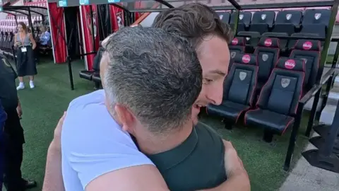 Tom Lockyer is embracing a paramedic, whose back is towards the camera. Tom's face can be seen over the man's right shoulder and he has his right arm wrapped around the man's back in a hug. Tom is wearing a white T-shirt and the paramedic is in his dark green uniform. They are standing on the edge of a pitch at a football stadium and the empty stand is behind them
