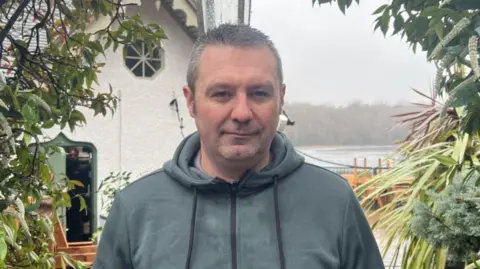 Sam Morgan is stood infront of the still standing part of the venue in a blue teal hoodie. 