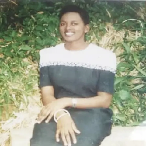 Emertha Uwanyingira Emertha as a teenager. She has cropped black hair and is wearing a black dress with a white section across the shoulders and chest, and a couple of bracelets on each arm. She is sitting on a wall with green plants behind her and smiling at the camera. 
