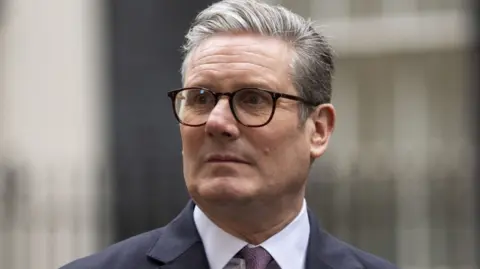 PA Media Headshot of Sir Keir Starmer. He stands outside what appears to be an out-of-focus Downing Street. He wears a dark jacket, a white shirt and a burgundy tie, with brown-rimmed spectacles and grey hair styled in a slight quiff.