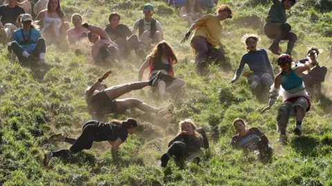 PA Media Competitors in a cheese rolling race kick up clouds of dust as they head downhill in Gloucestershire