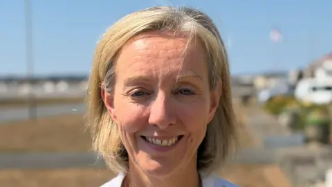 A woman called Ruth Thomason smiles at the camera while standing outside an office in Guernsey. She has fair hair and the top row of her teeth are showing.