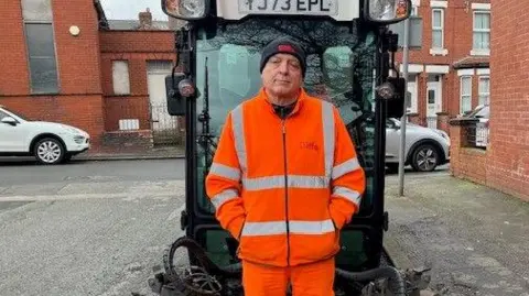 Man in his 60s stands on the pavement next to a clear-sided road sweeping vehicle with large yellow bristles on the front. He is wearing an orange high-visibility jacket and trousers and a black woolly hat.