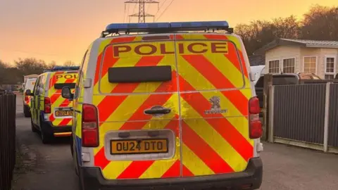 A rear view of three police vans lined up, one behind the other, on a concrete driveway, with a mobile home on the right.