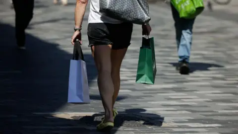 Woman wearing yellow strappy sandals walking down a High Street carrying two shopping bags, one in each hand