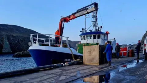 Tammy Hine A boat by a dock, with three people standing close to it. The boat is blue and white, with a man on it, and a crane moving a box off it. The dock is wet. Cliffs are to the left and a white van to the right. 