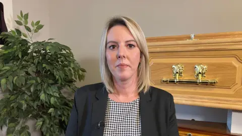 Carly Baker, MG Evans & Sons, Family Funeral Directors, is wearing a black blazer and chequered top and is stood in front of a light pine coffin with gold handles and a plant on the left of the image