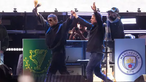 Getty Images Liam and Noel Gallagher are raising their hands to the audience as they walk on stage during Oasis's reunion tour