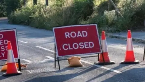 Red road closure sign in the middle of the road with a sandbag at the bottom to hold it in place  and cones in the road to stop motorists.