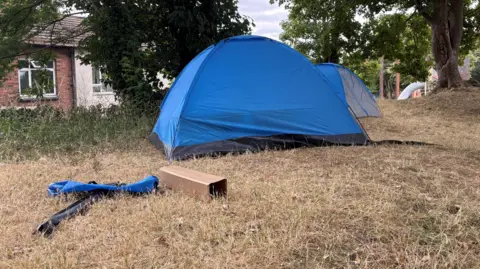 Ben Schofield/BBC Two tents pitched on grass. There are houses to the left and trees in the background.