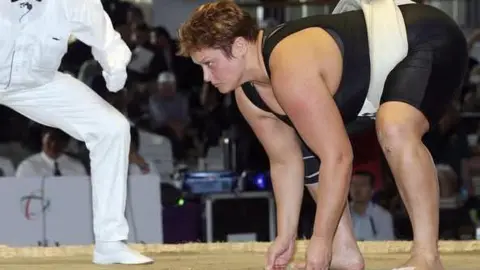 Adele Jones A sumo referee and Jones on a raised ring during a match. Jones is positioned in a low starting stance, with both hands touching the ground. She is wearing a black athletic outfit along with a traditional mawashi tied at the back. The referee, dressed entirely in white, is leaning forward as if preparing to signal the start of the bout. In the background, an audience is seated in a large indoor venue, watching the action unfold.