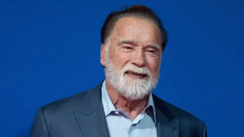 Arnold Schwarzenegger, an older man with black hair and a white beard standing smiling. He is wearing a blue shirt and dark jacket. The background is blue.
