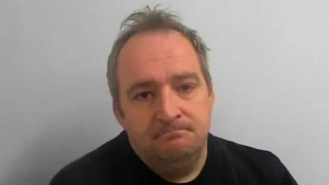 The photograph shows a man seated against a plain, light grey background, staring at the camera looking glum. He has short hair and is wearing a black crew-neck sweater.
