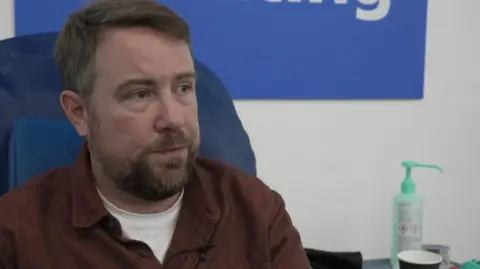 James Allen, wearing a brown shirt and white T-shirt is sitting in a blue hospital chair, with medical ephemera on a locker next to him.