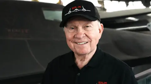 The head and shoulders of Buz Carpenter, standing in front of a Blackbird spy plane. He is wearing a black baseball cap and a black T-shirt with blackbird stitched on the left side. He is smiling broadly. 