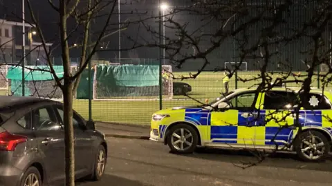 Un automóvil policial estacionado frente al campo deportivo. Se han cubierto goles de fútbol de cinco al lado de la lona verde que oscurece el sitio del accidente. Se puede ver que la parte delantera del auto negro sale detrás de uno de los objetivos. Los oficiales de policía se encuentran cerca.