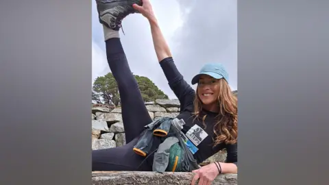 Handout Avril Langard-Tang poses during her walk with her leg stretched out while holding the bottom of her foot. She is smiling and wearing a light blue cap and mostly black clothes. 