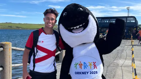 Steven Mackay is stood at the harbour in Orkney smiling to take a photo next to the games mascot, Orla the Orca.