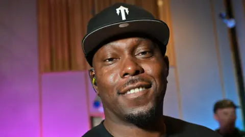 Close up photo of Dizzee Rascal wearing a black t-shirt and black cap with a white logo. He is smiling.