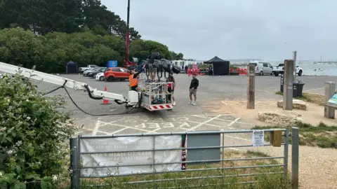 Newsquest Film production setup in a waterfront car park, featuring a camera crane, lighting equipment, crew members, and tents near a marina."