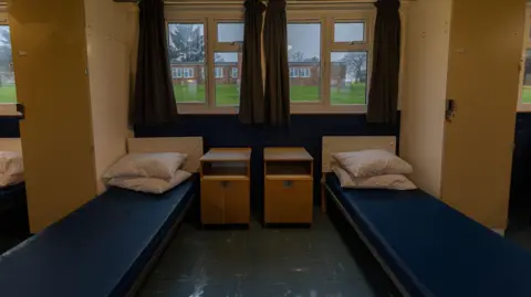 Home Office A room. There are two blue beds with white pillows. It looks basic and sparse. 