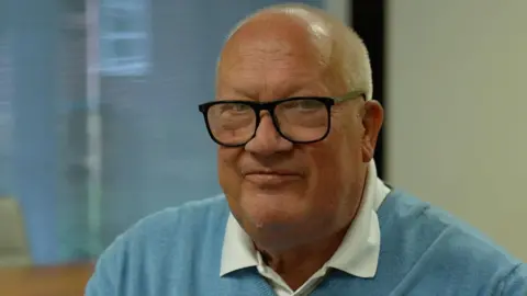 Rob Howard faces the camera wearing black framed glasses, a white shirt and light blue jumper.