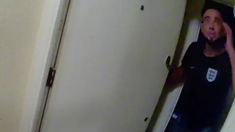 A man, Mohammed Durnion, who has short dark hair and a beard, is standing by a door with his hand to his head.