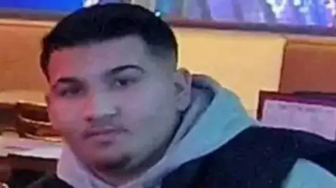 A young man with short black hair wearing a grey hoodie and black vest.