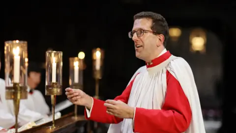 Submitted Daniel Hyde in white and red robes, conducts the choir. He is wearing glasses and has short, dark hair.