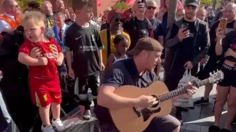Mark Phillip sits on a stone bench playing an acoustic guitar surrounded by a crowd of people, some recording on their phones. A little blonde-haired boy in a full replica Liverpool kit is being held by a woman behind him.