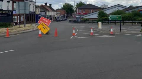 Town centre junction with road closed diversion signs and traffic cones blocking lanes, nearby shops including a pub and an Aldi supermarket visible in the background under cloudy skies