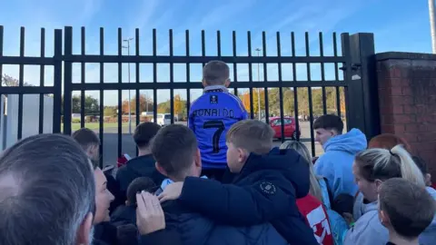 children outside the gate of St Mirren FC with one child in a blue Ronaldo shirt sitting on someone's shoulders