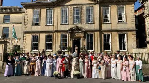 Around 50 people wearing Regency clothing standing outside Parade House in Trowbridge. It is a large historic stone building with 13 sash windows. It is a sunny day and everyone is lined up outside the entrance, ready for a photo.