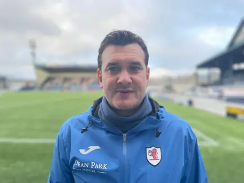Raith manager, Ian Murray - man looking at camera, wearing a blue jacket with the club logo with a football pitch in the background out of focus.
