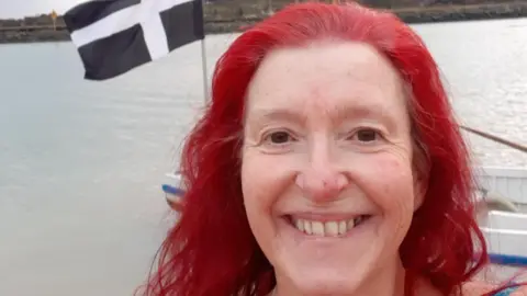 Louise Snelson takes a selfie in front of a Cornish flag on a boat in a harbour. She has dyed red hair and has a broad smile on her face.