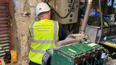 A man is fitting a meter in a stone shed. He is kneeling down next to a wall and there is a green generator behind him. 
