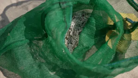 A dark green mesh bag filled with tiny white plastic pellets and sand. The bag is soft and flopped over so the majority of the pellets are only partly visible through the mesh.