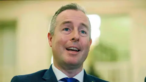 Paul Givan is wearing a navy blazer, white shirt and blue tie. He has short grey hair. The background is blurred. 