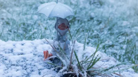 Becca C A smal duck statue wearing a white coat with a bow and holding an umbrella sits in long grass. There is snow lying on the ground.