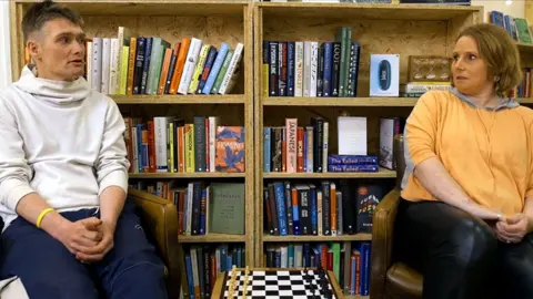 Colin MacMillan is wearing a white top and blue trousers. He is looking at his partner, Michelle, while they talk in front of a bookcase.