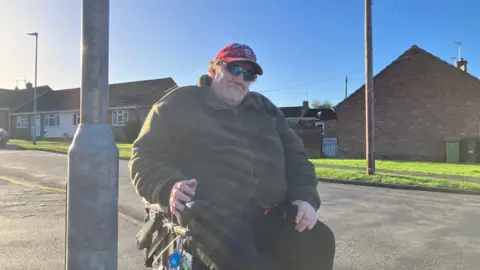 Andrew is in an electric wheelchair and is posing next to the lamppost. He wears a thick fleece and a red baseball cap.
