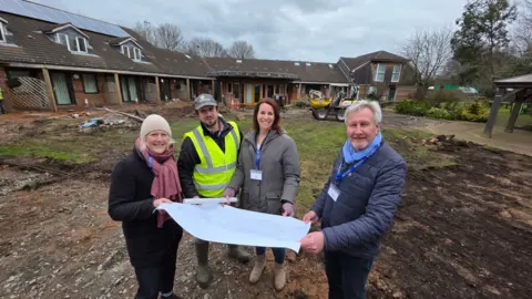 Acorns Children's Hospice Four people standing on a patch of grass that has been dug up, holding a large piece of white paper that has a large drawing on it. There is a red brick building in the background and the land they are standing on is a building site. There is a digger behind them.