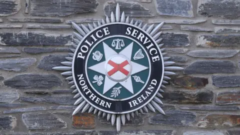 The picture shows the PSNI crest with the words Police Service of Northern Ireland on it. It also has a white star and red diagonal cross, as well as the symbol of the crown, the shamrock, a harp and scales of justioce