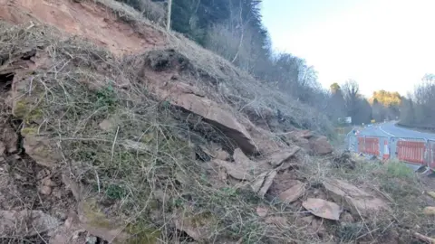 A slope of rocks near to a main road. The area has been cordoned off by gates and orange cones