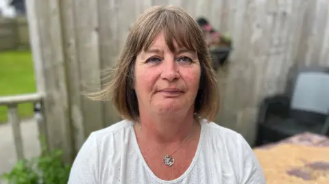 A woman in her 50s is sat on a garden bench with a fence behind her. She has mid length brown hair in a bob hair cut, and is wearing a white top with a silver necklace. She has a passive expression. 