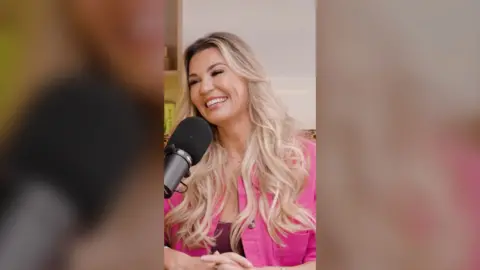 Christine McGuinness is smiling and wearing a pink jacket. She is sat in front of a mic and has her head tilted to the side.